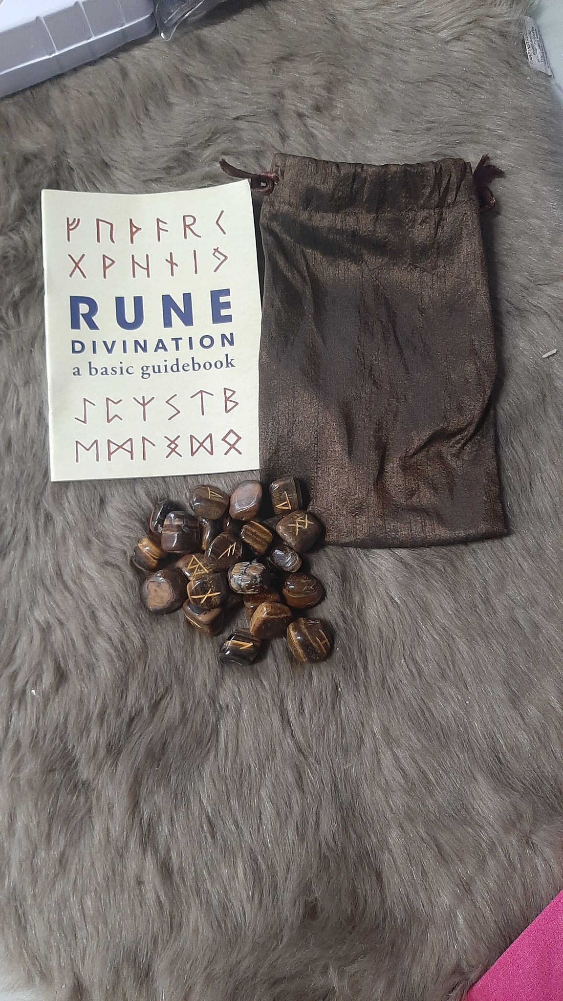 Set of dark stones, a small book titled 'Rune Divination', and a brown drawstring bag on a textured surface.